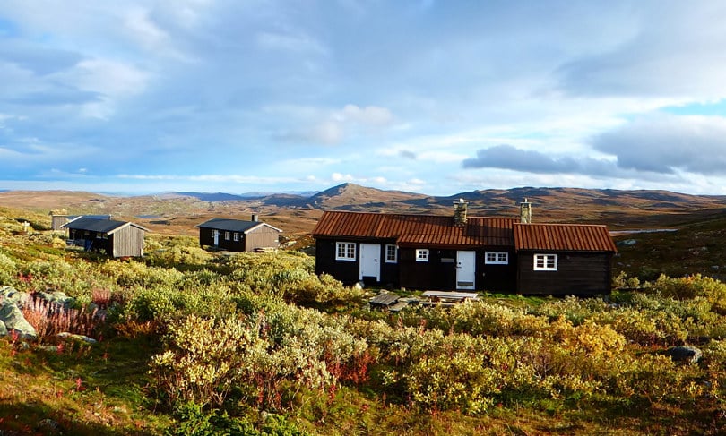 Tre brune hytter ved siden av hverandre i lav høstsol på fjellet.