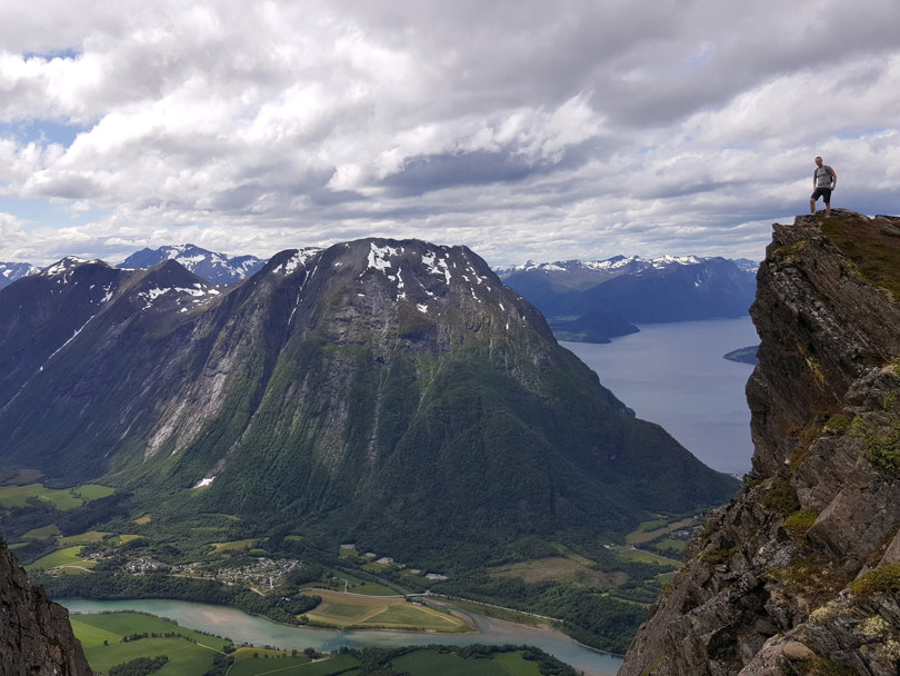 Mann i grå t-skjorte og svart shorts står ytterst på en klippeformasjon og ser utover en dal omgitt av høye fjell.