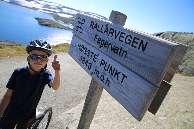 Gutt på sykkel holder tommelen opp ved siden av et skilt på fjellet.