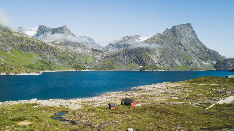 Rød hytte ved et stort fjellvann med høye fjell i bakgrunnen.