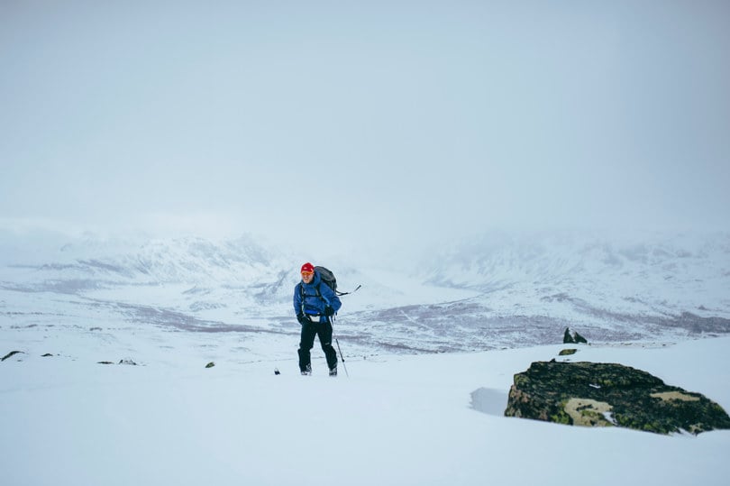 Skiløper i blå jakke og svart bukse går oppover ei fjellside i gråvær. 