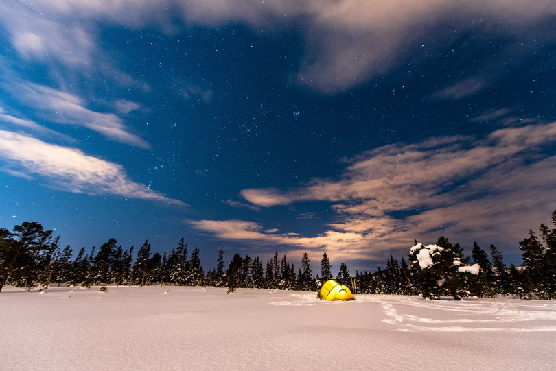 Lite telt med lys inni står på en åpen snødekt flate inntil en skog. Stjernene skinner på nattehimmelen. 