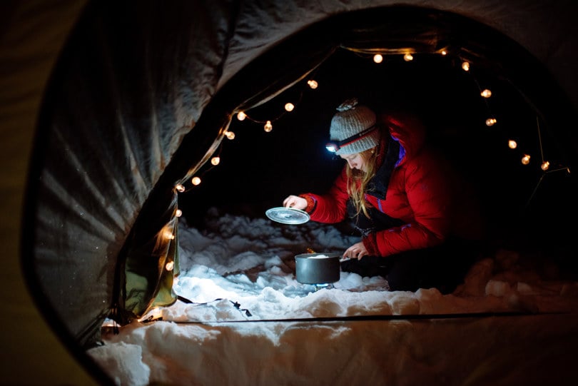 Person i rød dunjakke sitter på huk og lager mat utenfor en teltåpning. Personen har hodelykt som lyser opp foran. I teltåpningen henger en lyslenke.