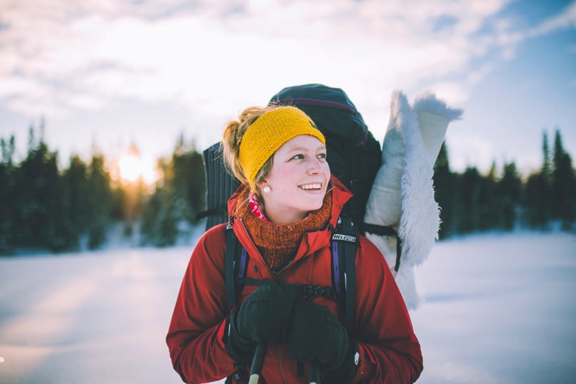 Kvinne i rød anorakk og gult pannebånd, lener seg på skistavene, ser til siden og smiler. På ryggen har hun en stor sekk.sekk med 