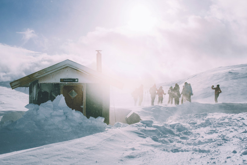 Flere personer står utendørs i snøføyke ved siden av ei lita hytte.