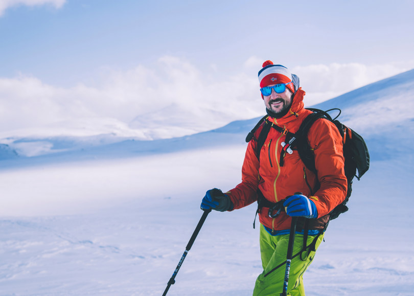 Person i oransje jakke og grønn bukse, med lue og solbriller, lener seg på skistavene og smiler.