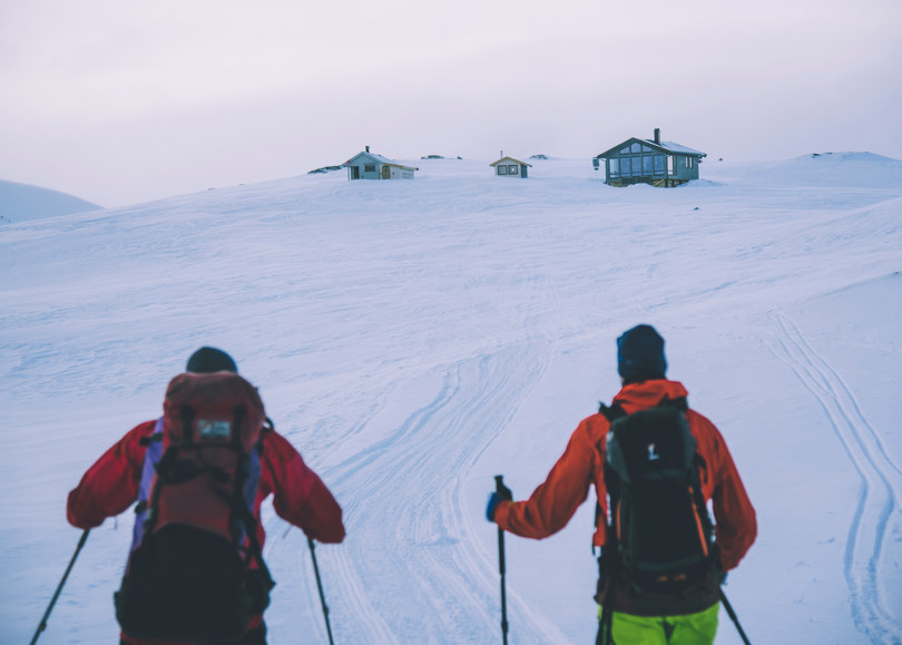 To personer med ryggsekk går på ski oppover mot tre hytter i ei snødekt fjellside.
