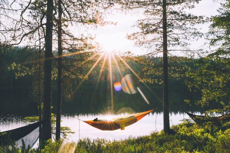 Person ligger i en gul hengekøye mellom to trær rett ved et skogsvann. Sola står opp over åsen i bakgrunnen.