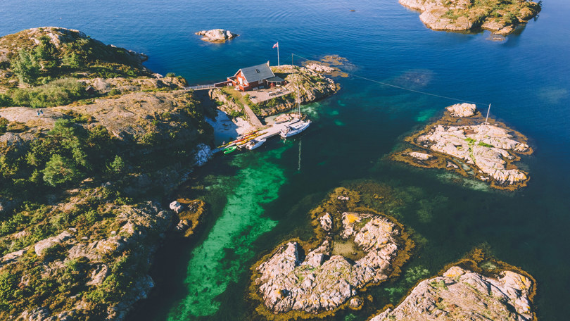 Rød hytte ytterst på en holme ved sjøen. Foran hytta ligger flere kajakker og båter ved ei brygge.