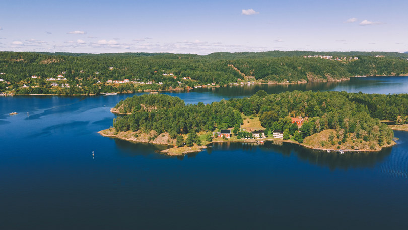 Fugleperspektiv på lang avstand av en liten skogkledd øy. Ved strandkanten ligger tre store og gamle hus.