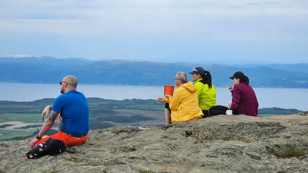 Fire personer på tur sitter på en fjelltopp med ryggen til kamera, og ser utover landskapet. Fjord synlig i bakgrunnen