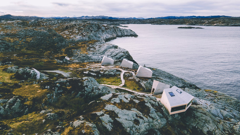 Dronefoto av fem små hytter ytterst på en naken holme ved havet.