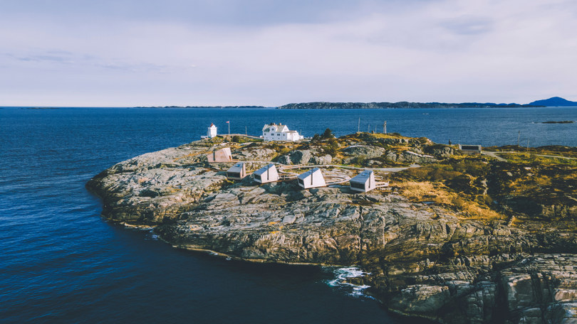 Dronefoto av fem små hytter og et fyr ytterst på en naken holme i havgapet.