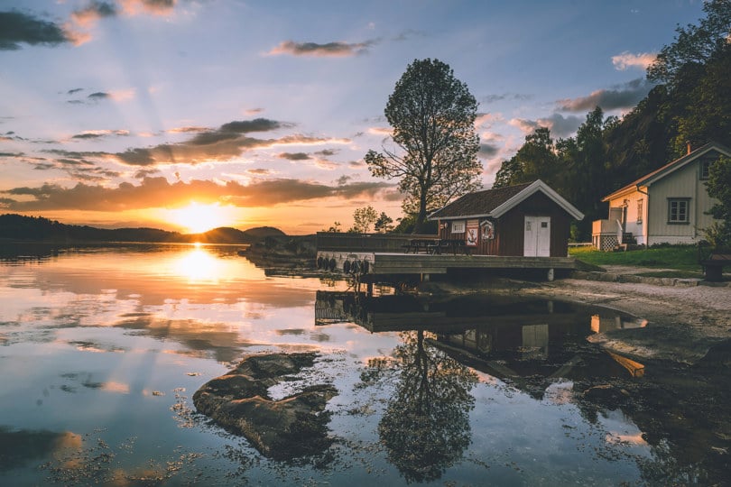 Rødt naust med brygge som stikker ut over vannet ved siden av en liten strand. Bak naustet ligger en liten hvit hytte. Sola går ned i bakgrunnen og lager et stemningsfylt lys.