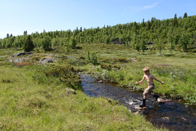 Gutt i shorts og støvler hopper over en bekk. I bakgrunnen skimtes ei hytte i en skogkledd ås.