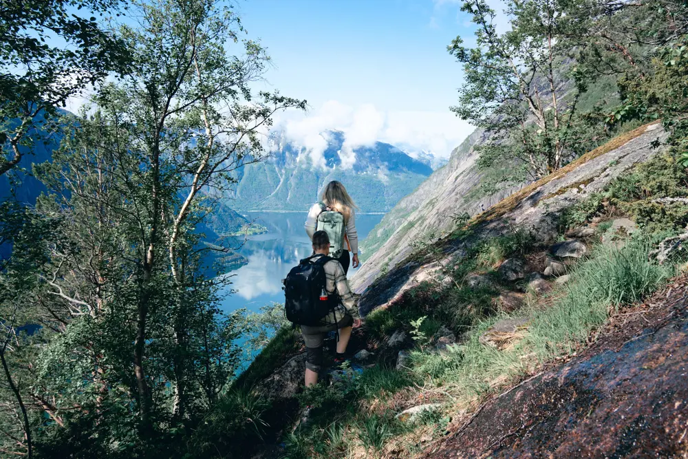 To personer går i skråning med fjord og fjell foran seg.