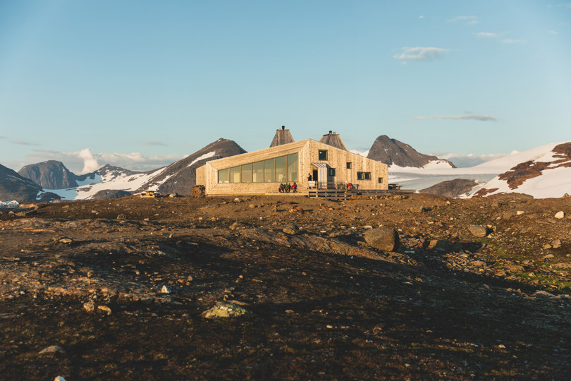 Grå og plankekledd hytte med store vinduer på snaufjellet. Utenfor hytta sitter flere mennesker. I bakgrunnen er det høye, spisse fjell og isbreer.