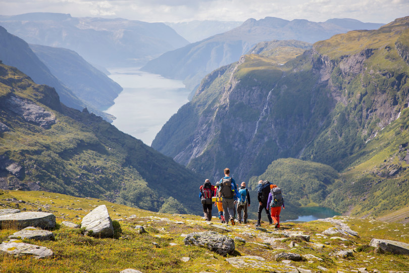 Turfølge med ryggen til går nedover en fjellside, mot en fjord omgitt av høye fjell.