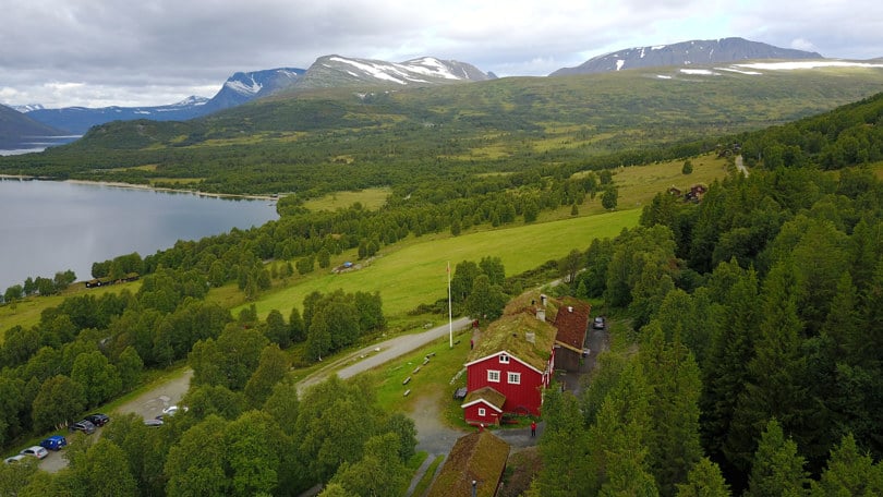 Fuglefoto av en rød hytte med torvtak. I bakgrunnen skimtes en sjø og høye fjell.