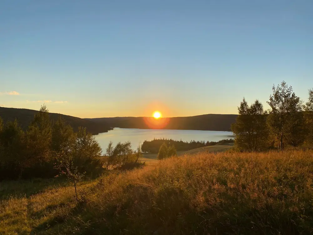 Et naturlandskap, med en eng i forgrunnen, en innsjø i mellomgrunnen, og en solnedgang på horisonten midt i bildet. Skyfri himmel. Trær på venstre og høyre side. 