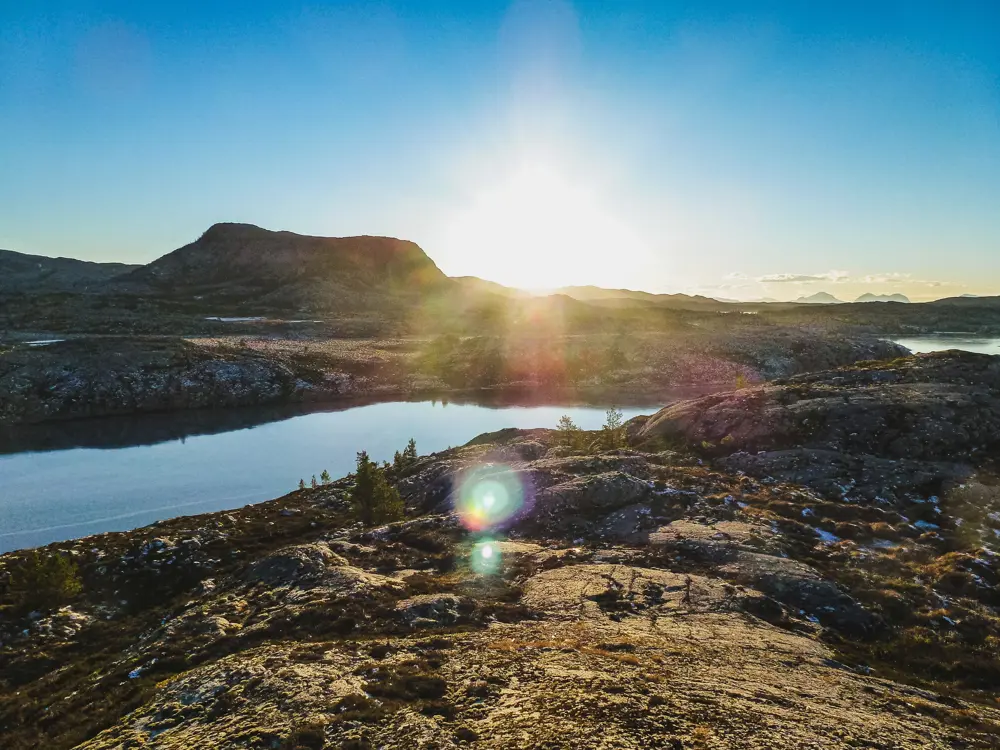 Åpent og steinete landskap delt av vann mellom forgrunn og bakgrunn. Et lavt og bredt fjell står foran horisonten midt i bildet, med solen rett til høyre for seg. Noen få spredte små planter her og der. En rekke fjell strekker seg ut lenger og lenger unna i det fjerne. 