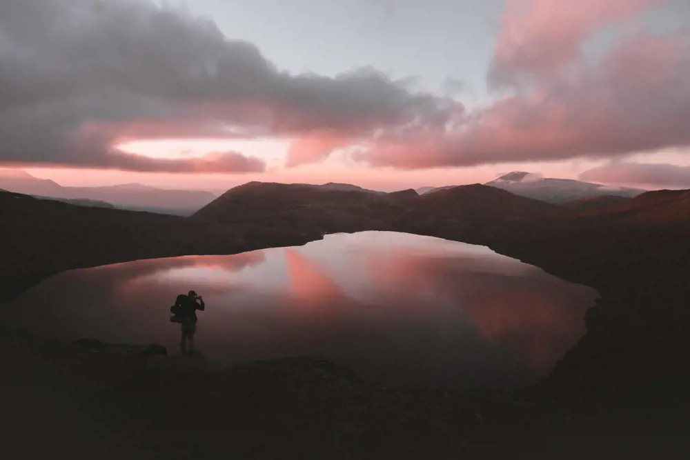 En speilblank innsjø, med en mann stående foran med et kamera, klar til å fotografere. Bak og rundt innsjøen er det lave fjell, og lave skyer dekker halve himmelen. Hele landskapet har et rosa-lilla lys over seg. 