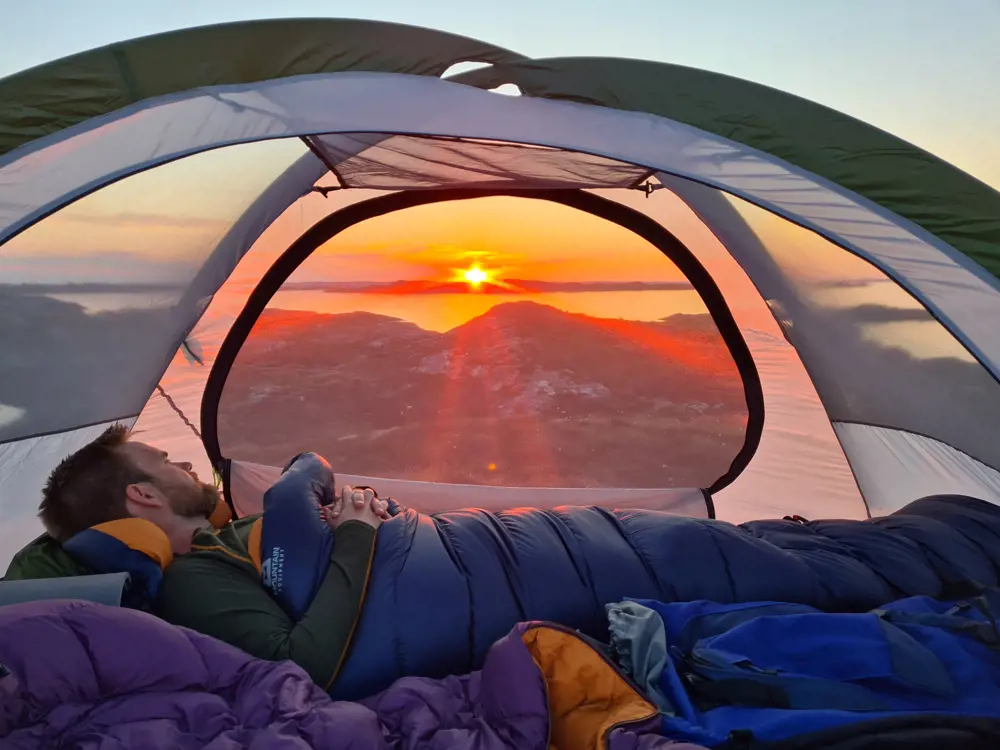Bilde av innsiden av et telt, hvor en mann ligger i sovepose og ser ut gjennom bakveggen av teltet, hvor en solnedgang avgir sterkt oransje lys. 