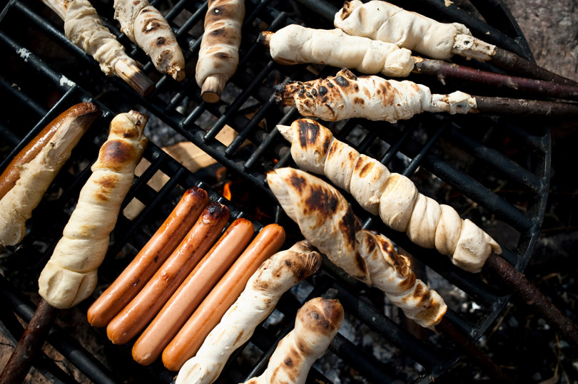 Pinnebrød og pølser i kryss og tvers på en grillrist over et bål.