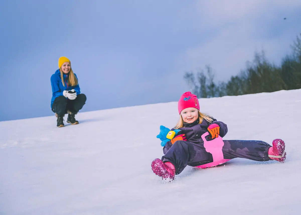 Jente på et rosa romeakebrett aker mot fotografen, mens en kvinne med langt blondt hår står på huk bak og ser på mens hun smiler. Snøkledd bakke, noen få trær i bakgrunnen til høyre. 