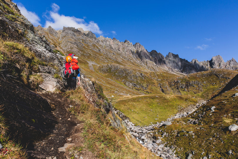 Person med rød sekk går langs en sti en bratt fjellside mot en dal omgitt av spisse tinder.