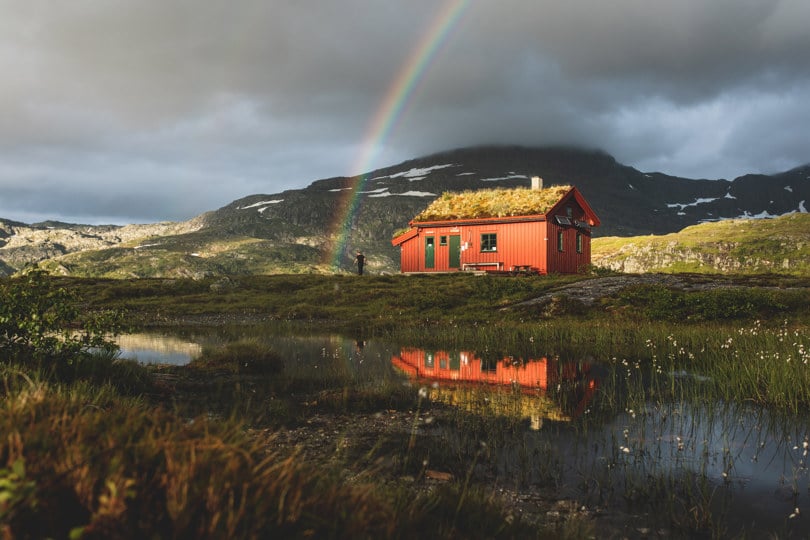 Rød hytte ved et lite vann på fjellet. På himmelen bak hytta er det en regnbue.