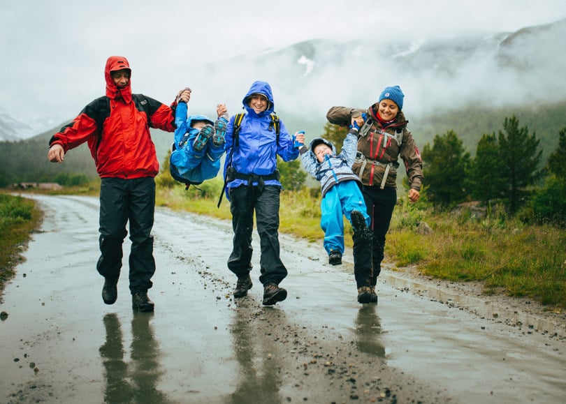 Tre voksne personer i regntøy løfter og svinger to barn mellom seg mens de går bortover en grusvei.