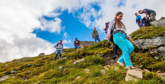 En gjeng med unge voksne på tur i fjellet. Helt foran ser du en ung jente med turkise bukser som smiler og går ned en skråning.
