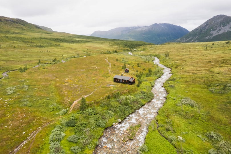 Fugleperspektiv av ei hytte som ligger ved en liten elv i åpent og grønt fjellterreng.