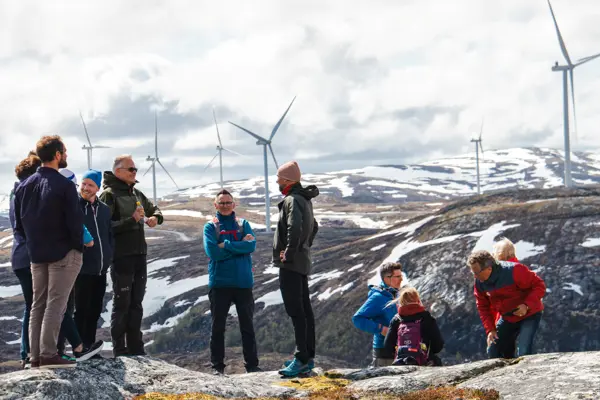 En gruppe med folk, kledd for kald vind, står oppstilt på en høy bakketopp og prater sammen, med en vindmøllepark synlig i bakgrunnen.