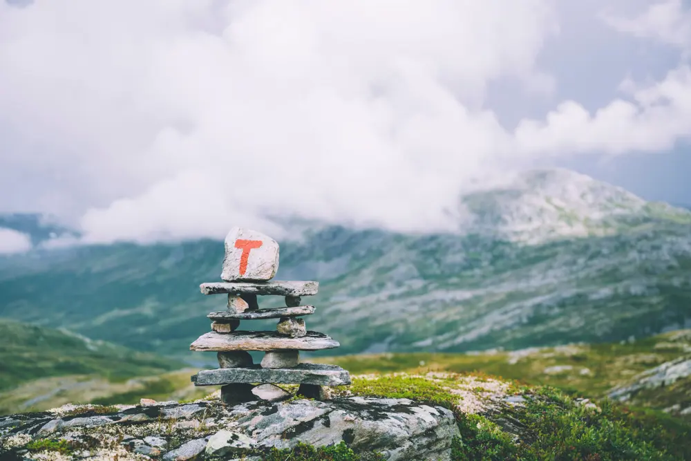 En varde laget av steiner stablet. Den øverste steinen har en rødmalt T på seg. fjellandskap i bakgrunnen.