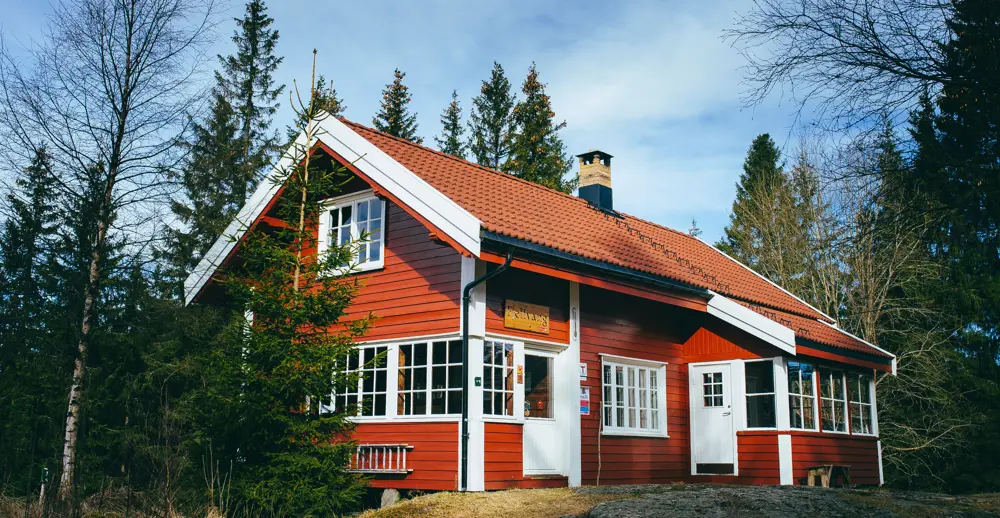 Rødmalt hytte med hvite karmer og oransje takstein i barskog. 