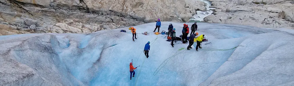 En gruppe mennesker klatrer oppover en blå isbre. Mye grå stein og en liten foss i bakgrunnen.