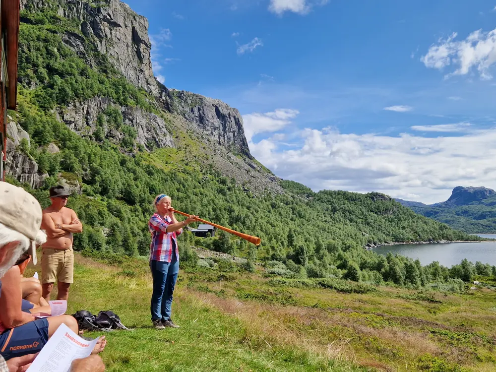 En kvinne står og blåser i et langt horn i et storslått fjelllanskap. Mennesker sitter i gresset og hører på henne.