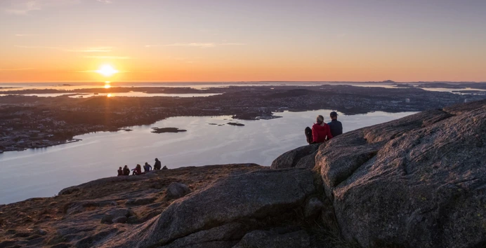 To mennesker sitter i solnedgangen på en fjelltopp og ser utover fjorden.