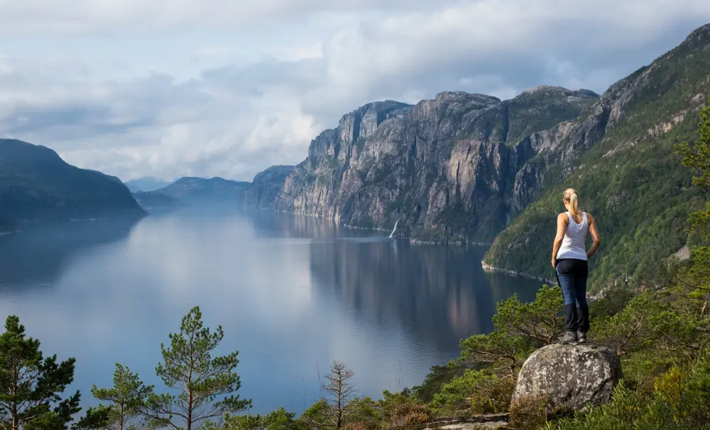 En dame med lys hestehale, iført hvit singlet og blå bukse står på et fjell og ser utover fjorden. 