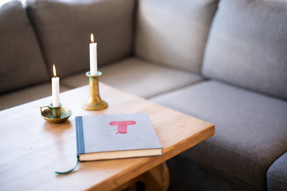 På et trebord foran en grå sofa står det to stearinlys som er tenkt, og det ligger en bok med rød T på forsiden. 