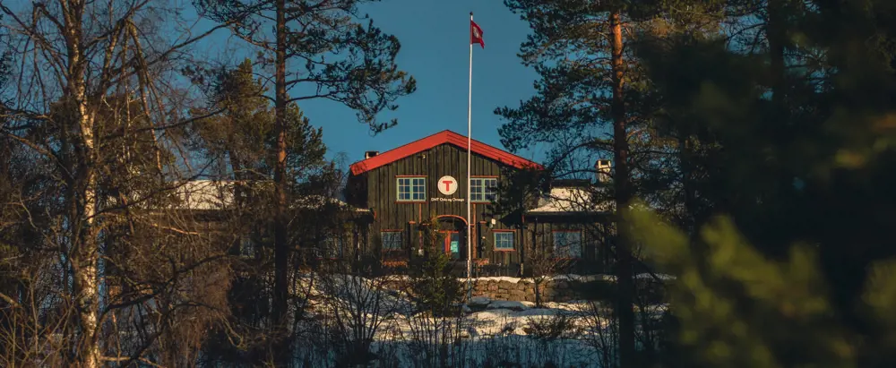 Svartmalt hytte med røde karmer og en flaggstang med norsk flagg