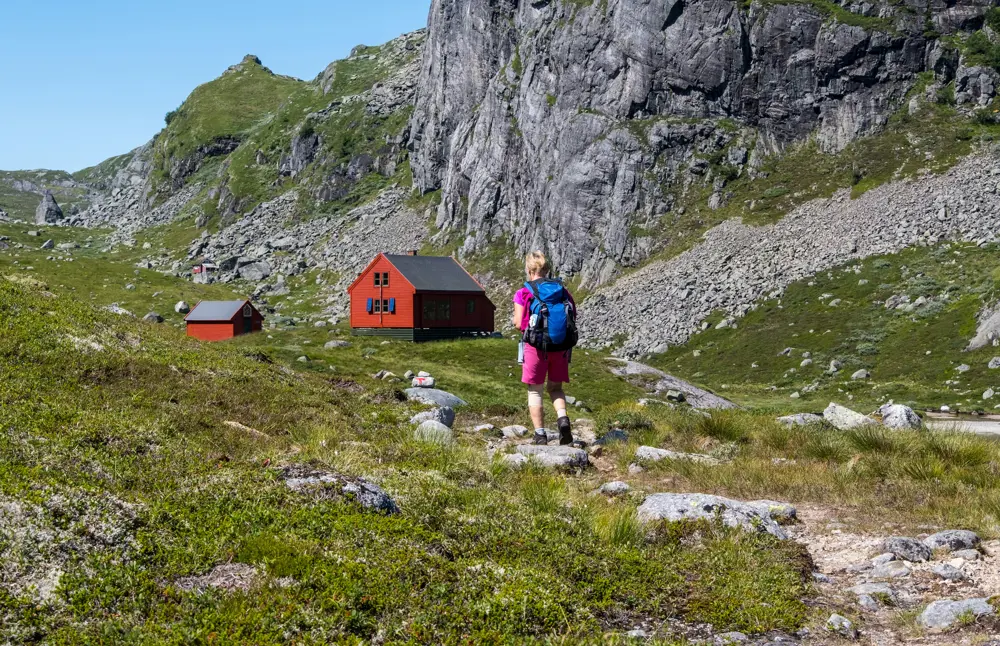 En kvinne i rosa turklær og blå sekk går mot en rød hytte i grønt terreng.