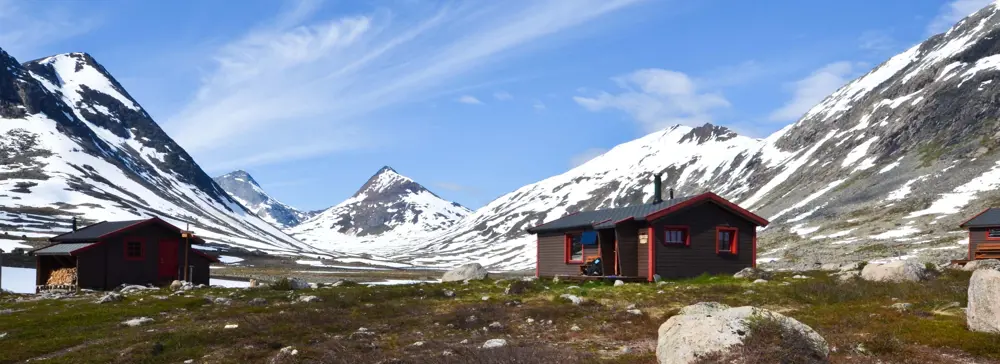 To hytter i sommerfjellet, snødekte fjell i bakgrunnen