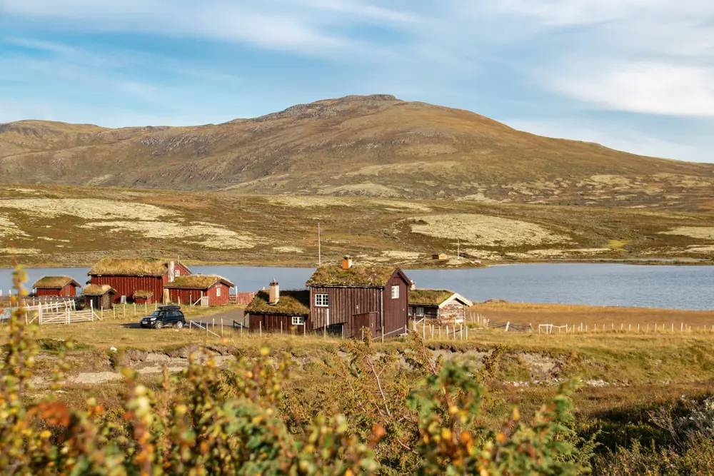 Bildet viser en liten hytte ved siden av en fjellseter i relativt flatt fjellterreng, et fjell og en stor innsjø i bakgrunnen. Fint vær med sol, og antydning til høstfarger på vegetasjon.