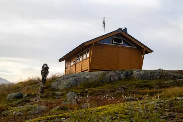 Gul hytte som ligger på en høyde. En mann går nedover fra hytta med sekk på ryggen.
