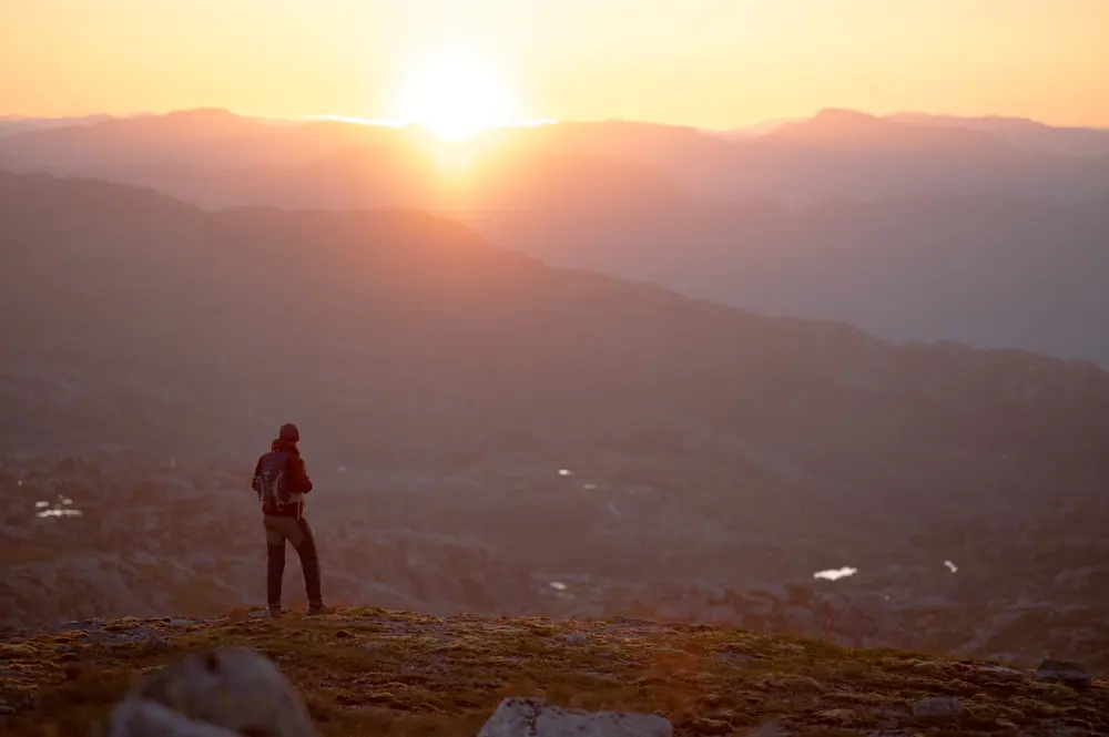 En person speider utover fjelllandskapet med sola som går ned bak fjellene. Oransje lys i bildet.