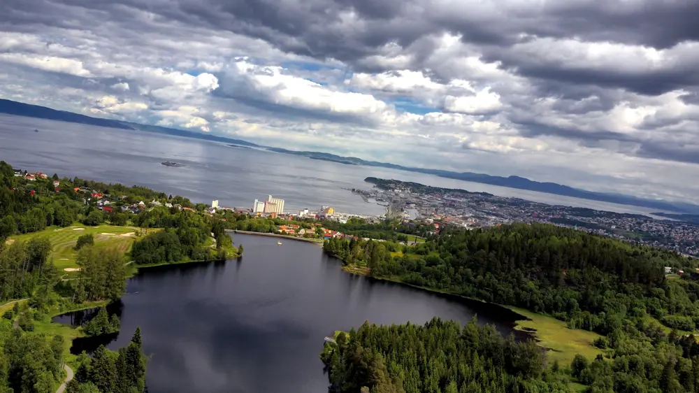 Dronebilde av Theisendammen sett ovenfra. En innsjø i bymarka, med fjorden synlig i bakgrunnen. Skyfull himmel.