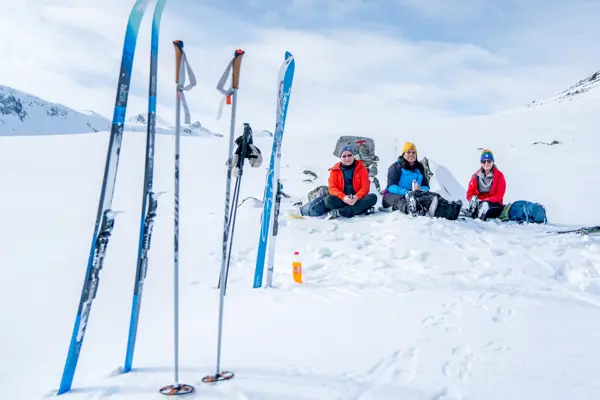 Tre personer i varme turklær sitter ved siden av hverandre i snøen, mens skiene og stavene deres er parkert stikkende opp av snøen foran dem.